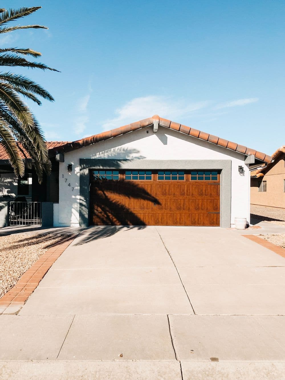 House with a tiled roof and wooden garage door, surrounded by palm trees and clear blue sky.